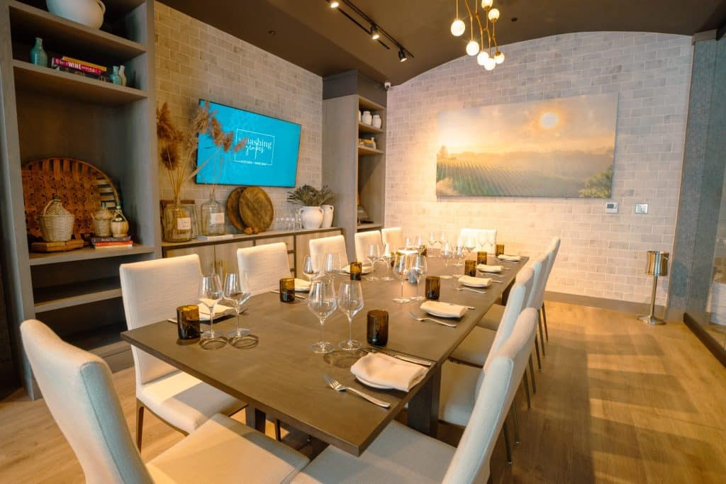 Dining room with a long wooden table set for a meal, white upholstered chairs, and wine glasses ready for guests in a modern restaurant setting.