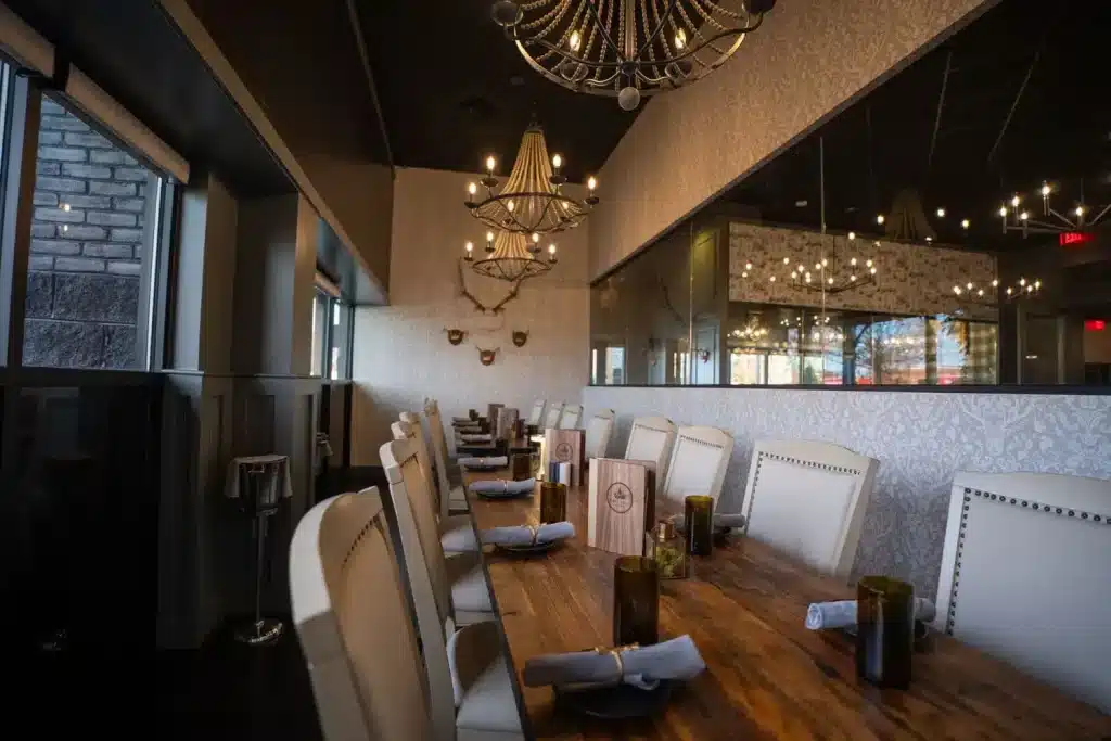 Long wooden dining table with white upholstered chairs in a chic restaurant, illuminated by crystal chandeliers and decorative wall patterns, set with napkins and menus.