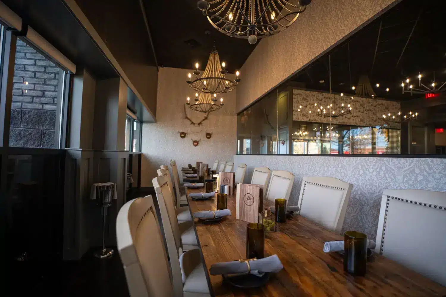 Long wooden dining table with white upholstered chairs in a chic restaurant, illuminated by crystal chandeliers and decorative wall patterns, set with napkins and menus.