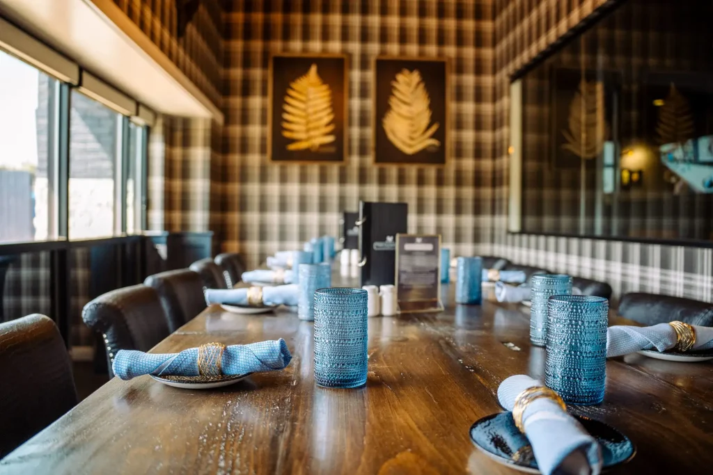 A long wooden dining table set with blue napkins and teal textured glasses in a plaid-walled restaurant interior.